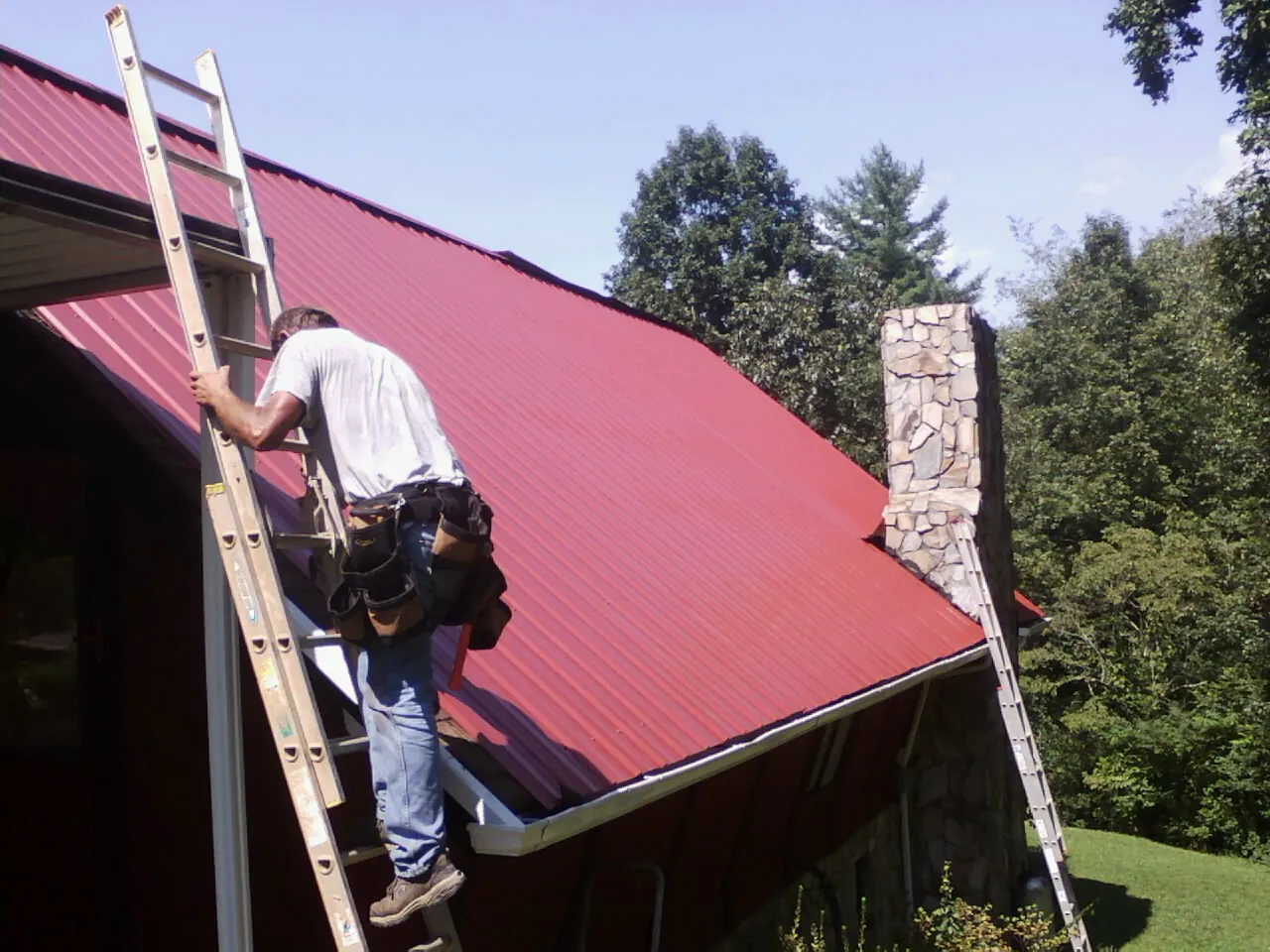 Roofer on a ladder beside a red metal roof and stone chimney — Soffit & Fascia Repair by Roofing Brothers in Boyce